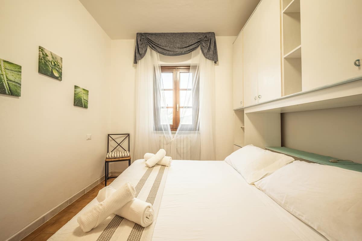Een net opgemaakt bed met witte lakens en een kussen met strepen, een stoel met een kussen, een raam met dunne gordijnen en een muur met drie ingekaderde foto's.