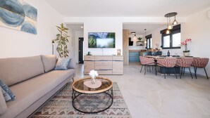 Ein modernes Wohnzimmer mit einer Couch, einem runden Couchtisch und einem Fernseher. Es gibt einen Essbereich mit rosa Stühlen und einem Holztisch.