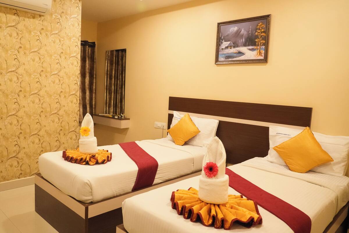 Quarto de hotel com duas camas, cada uma com colcha branca, almofadas decorativas vermelhas e amarelas, e uma toalha com uma flor amarela.
