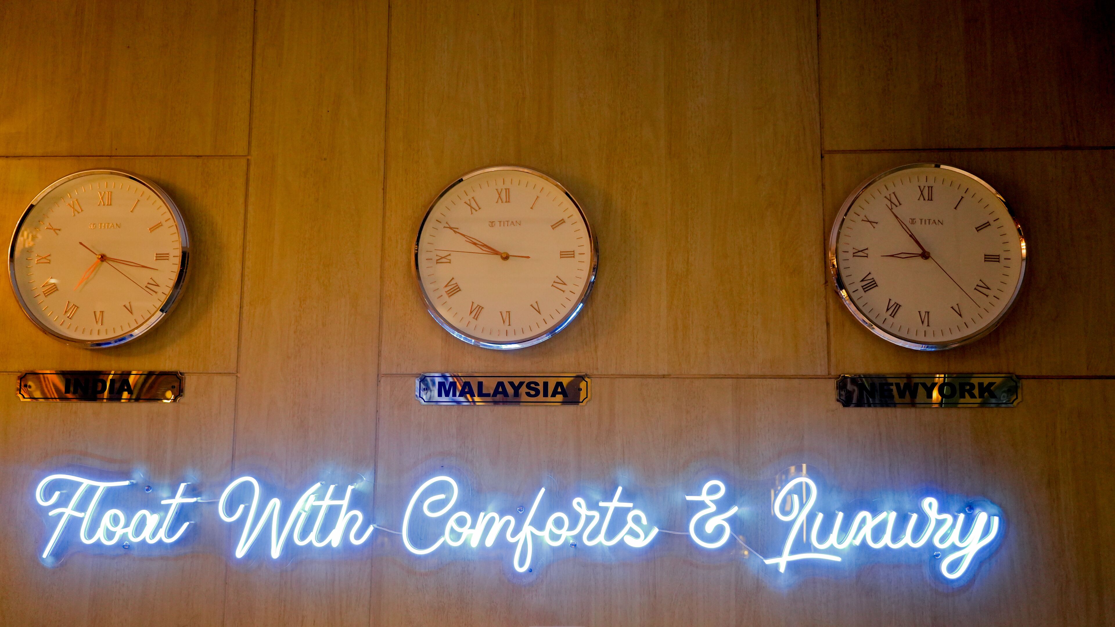Three clocks labeled with country names: India, Malaysia, and New York. Below the clocks, a neon sign reads "Float With Comforts & Luxury".