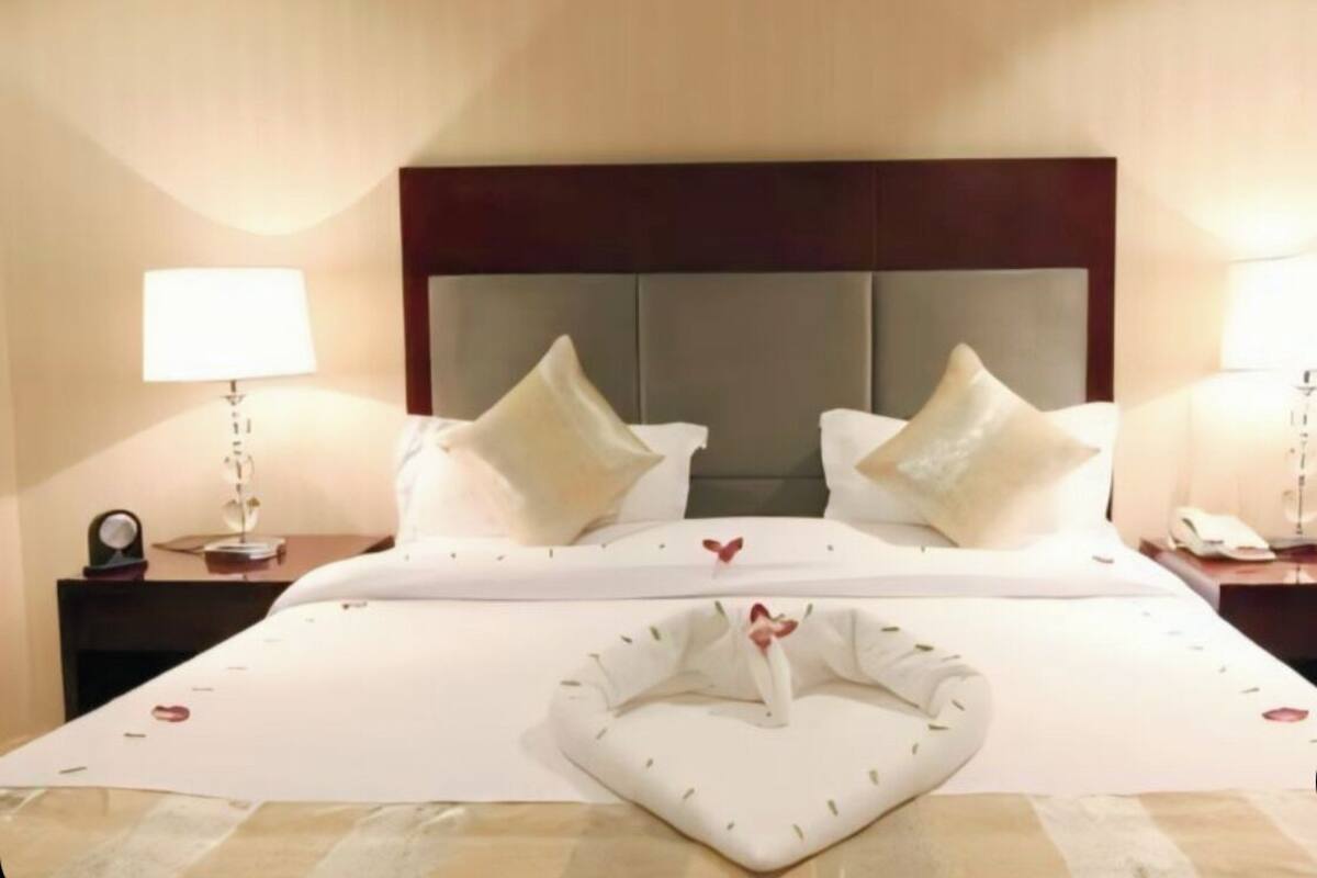 Quarto de hotel com uma cama grande, duas mesas de cabeceira, um criado-mudo, uma luminária e uma almofada decorativa em formato de cisne na cama.
