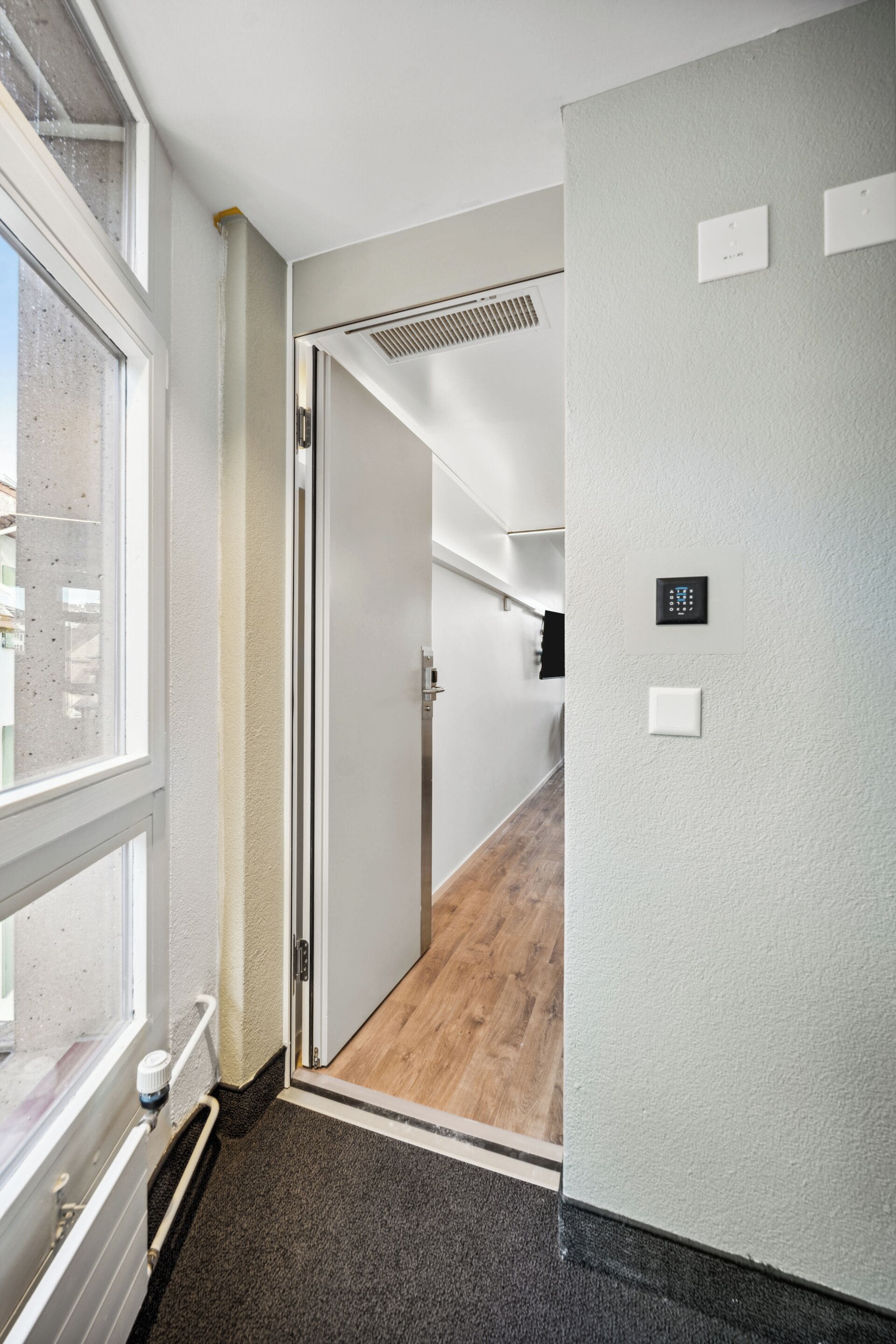 Un pasillo con una puerta que da a una habitación, una ventana y un termostato en la pared.