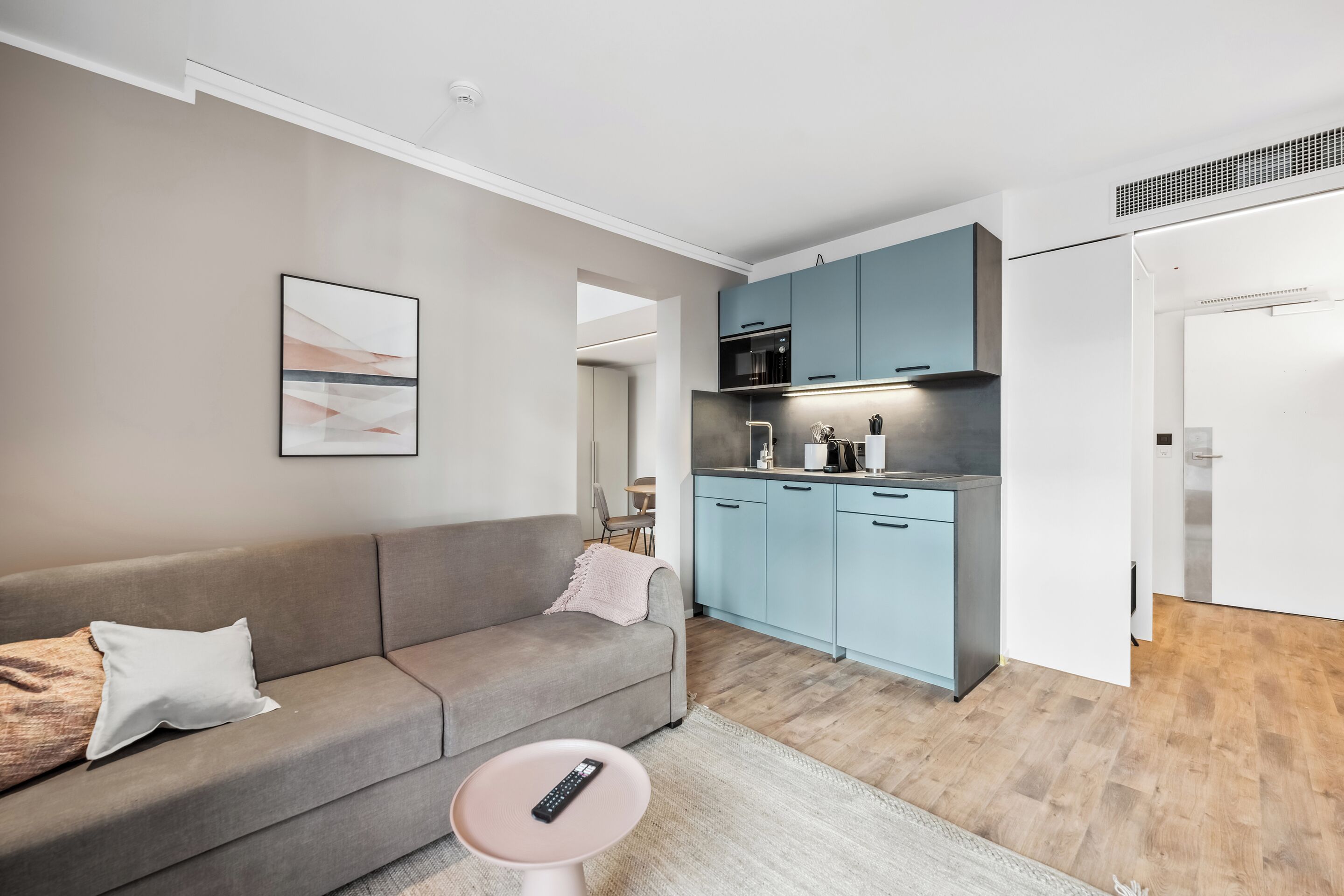 Una sala de estar moderna con un sofá gris, un control remoto sobre una mesita y una cocina con armarios azul claro y un microondas.