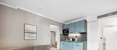 Una sala de estar moderna con un sofá gris, un control remoto sobre una mesita y una cocina con armarios azul claro y un microondas.