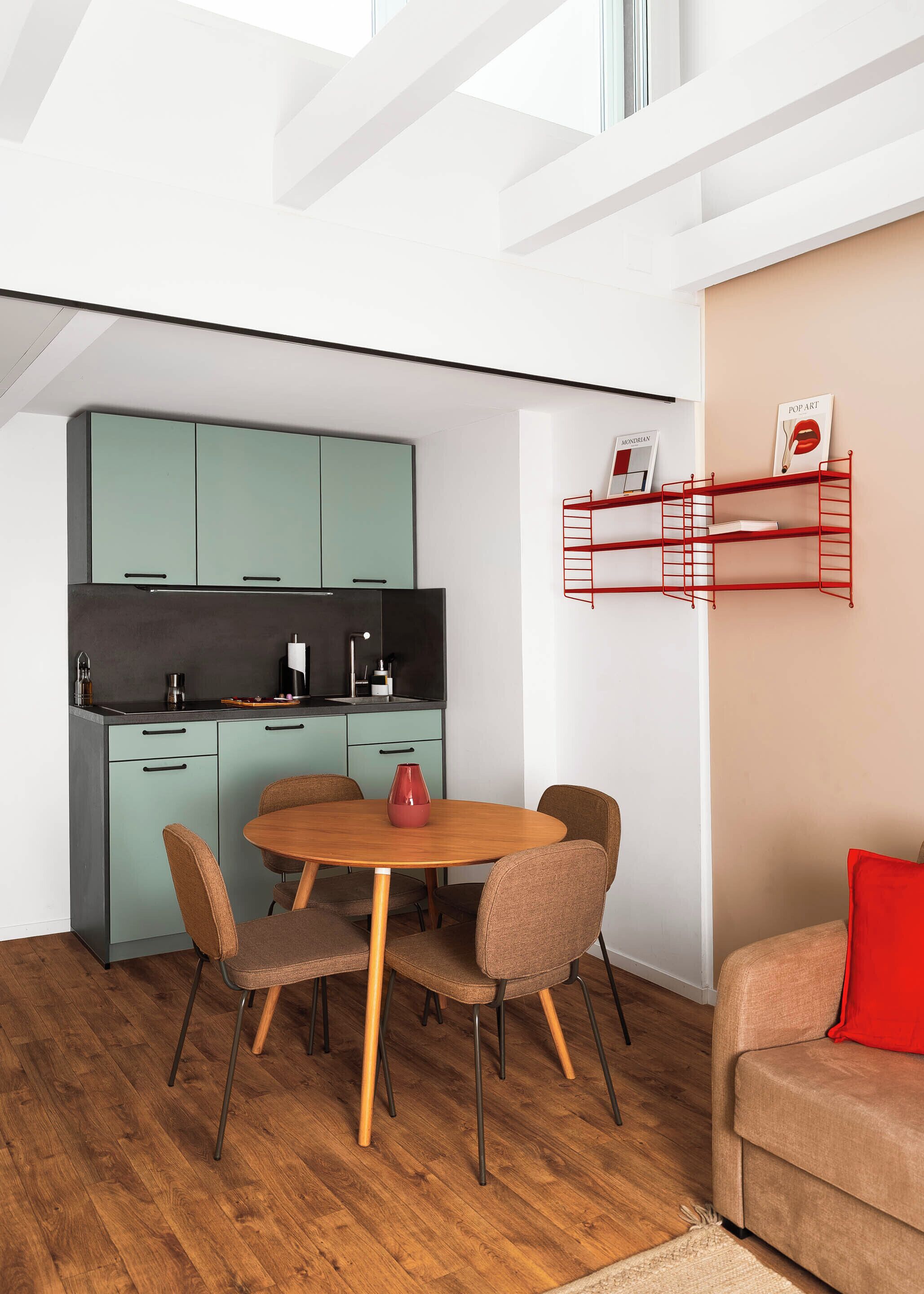 A modern kitchen with teal cabinets, a dining table with chairs, and a red shelf with books.