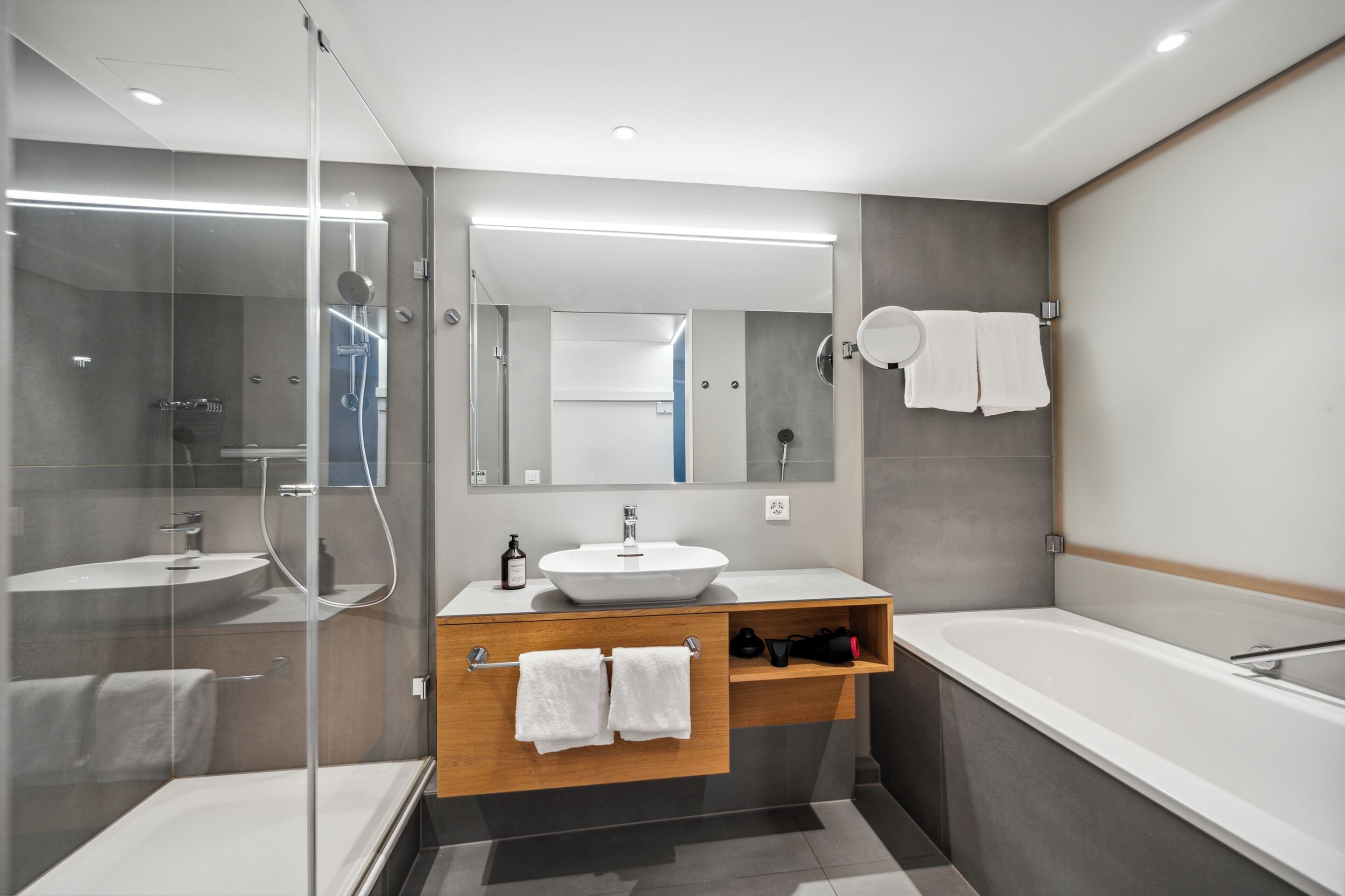 Un baño moderno con ducha de vidrio, tocador de madera con lavamanos y una zona de bañera separada.