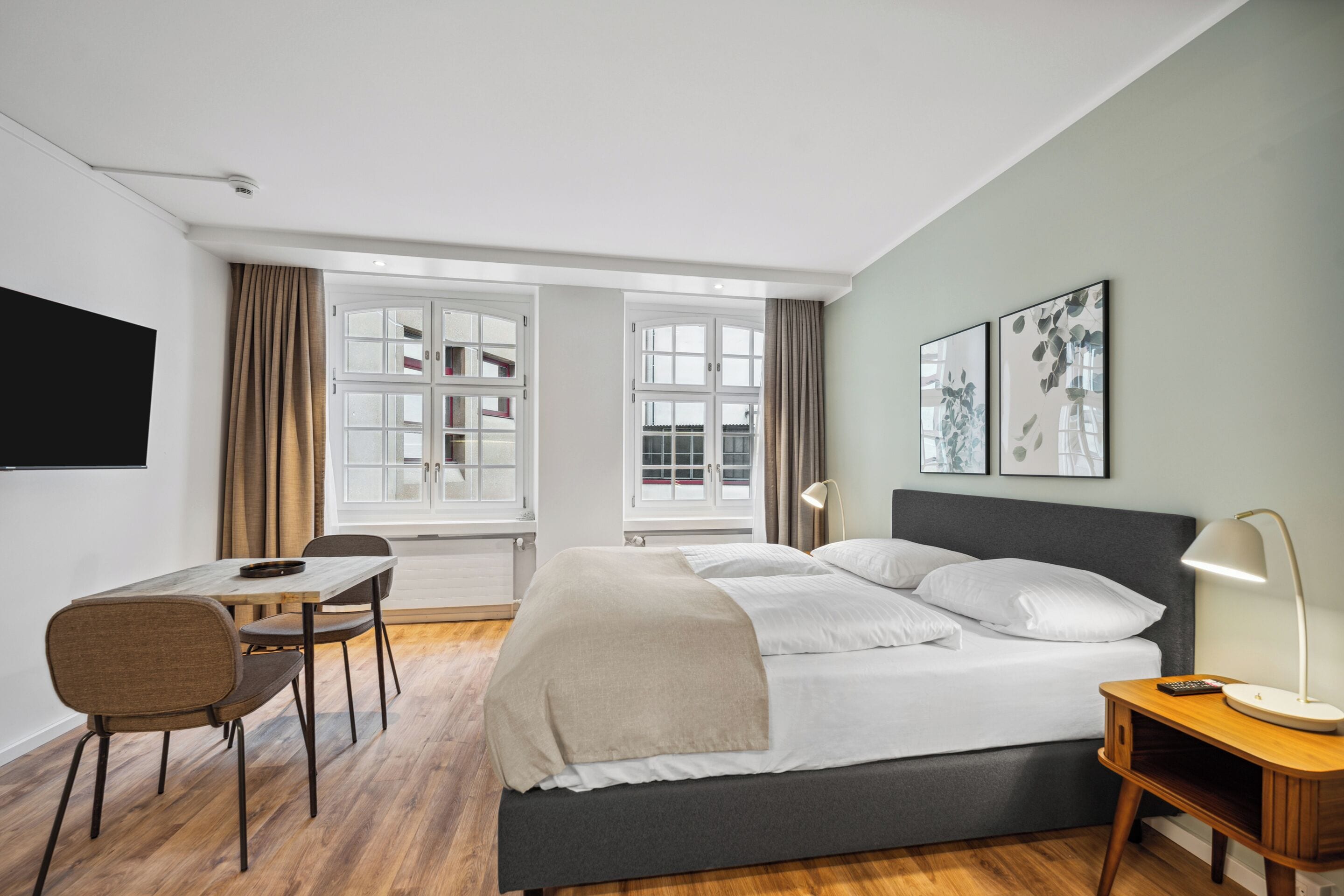 Una habitación de hotel moderna con una cama, un escritorio y una silla. Hay ventanas con cortinas, un televisor de pantalla plana montado en la pared y obras de arte en la pared.