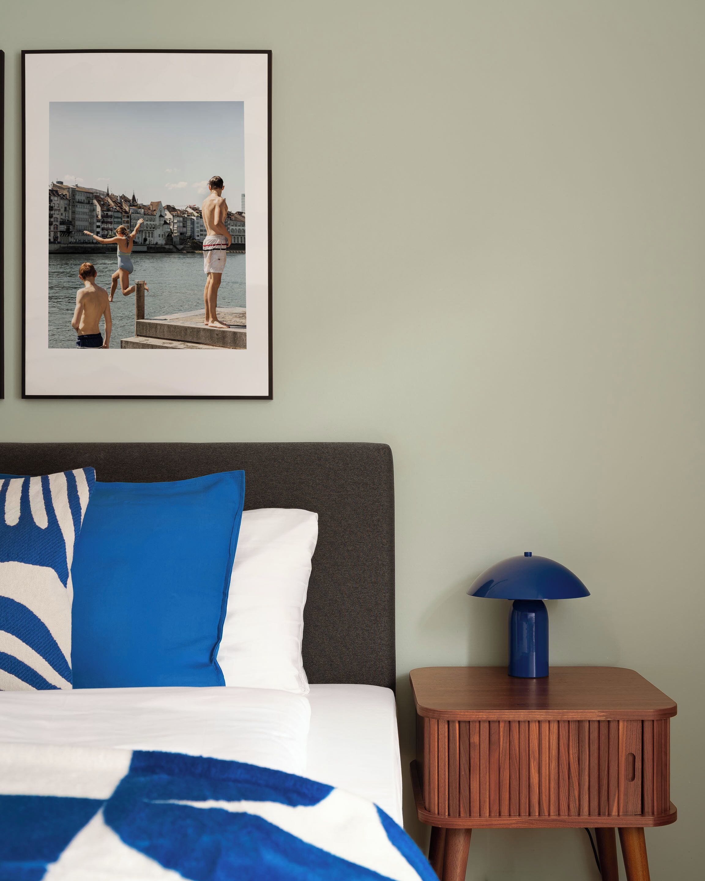 Un dormitorio con cama, almohadas, una mesita de noche con lámpara y un cuadro enmarcado en la pared.