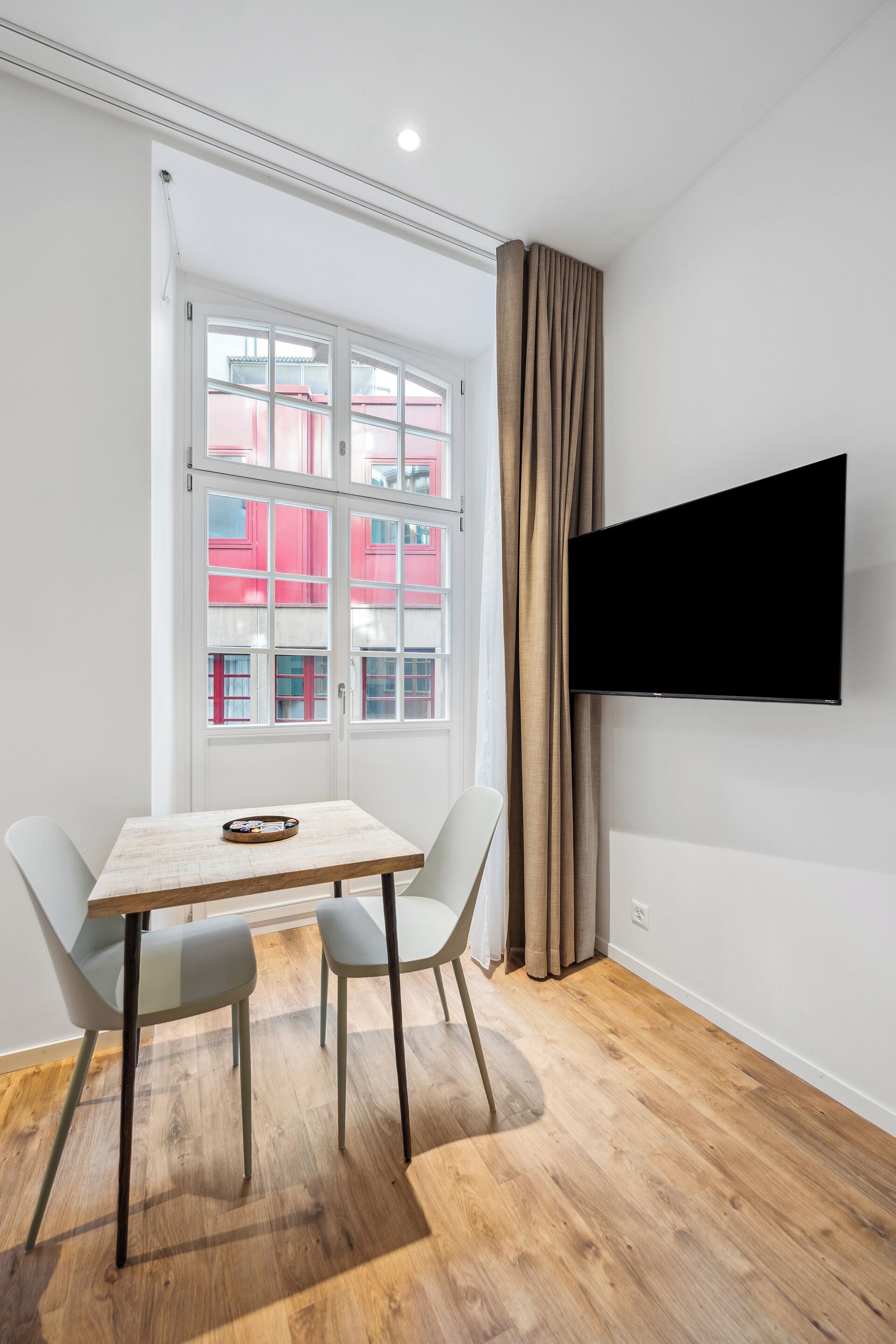 Una habitación moderna con suelo de madera, una mesa con sillas, un televisor de pantalla plana montado en la pared y una ventana con cortinas.