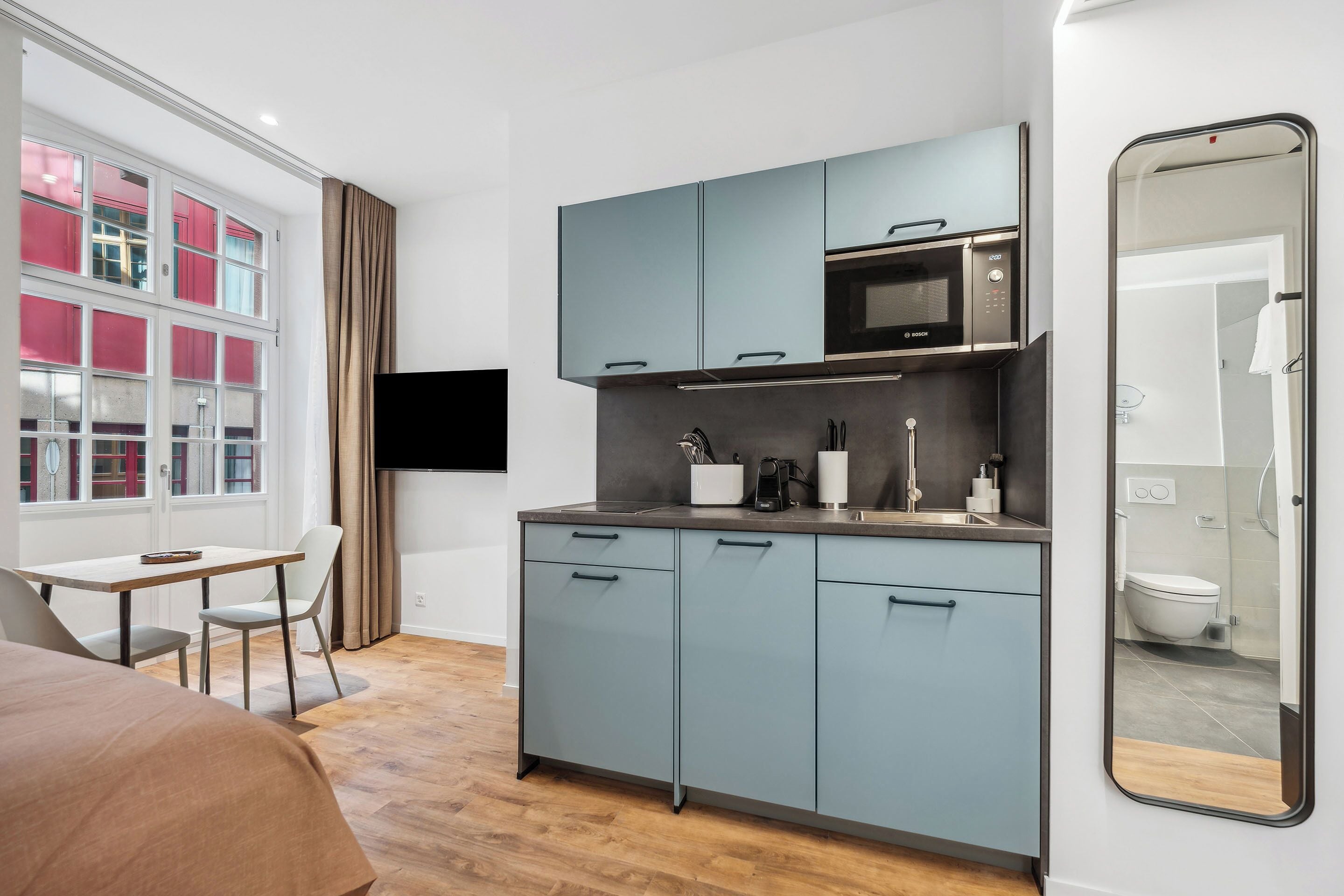 Habitación de hotel compacta con kitchenette, zona de comedor con mesa y sillas, ventana grande con cortinas y un espejo que refleja el baño.