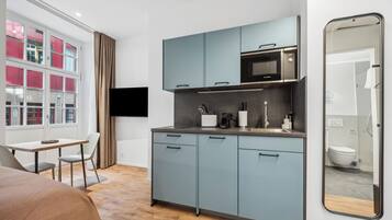 Habitación de hotel compacta con kitchenette, zona de comedor con mesa y sillas, ventana grande con cortinas y un espejo que refleja el baño.