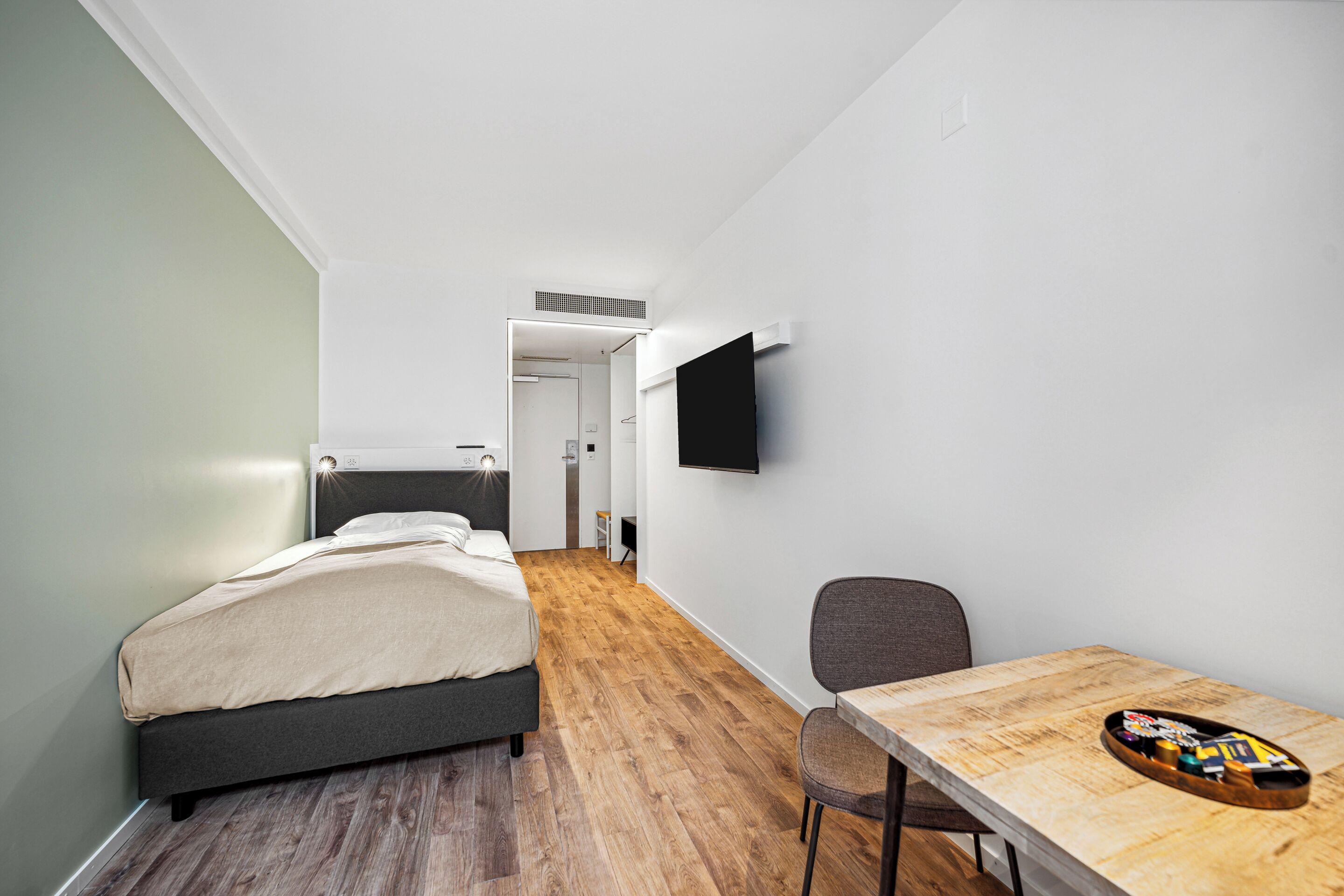 A modern hotel room with a bed, a desk, and a flat-screen TV mounted on the wall.