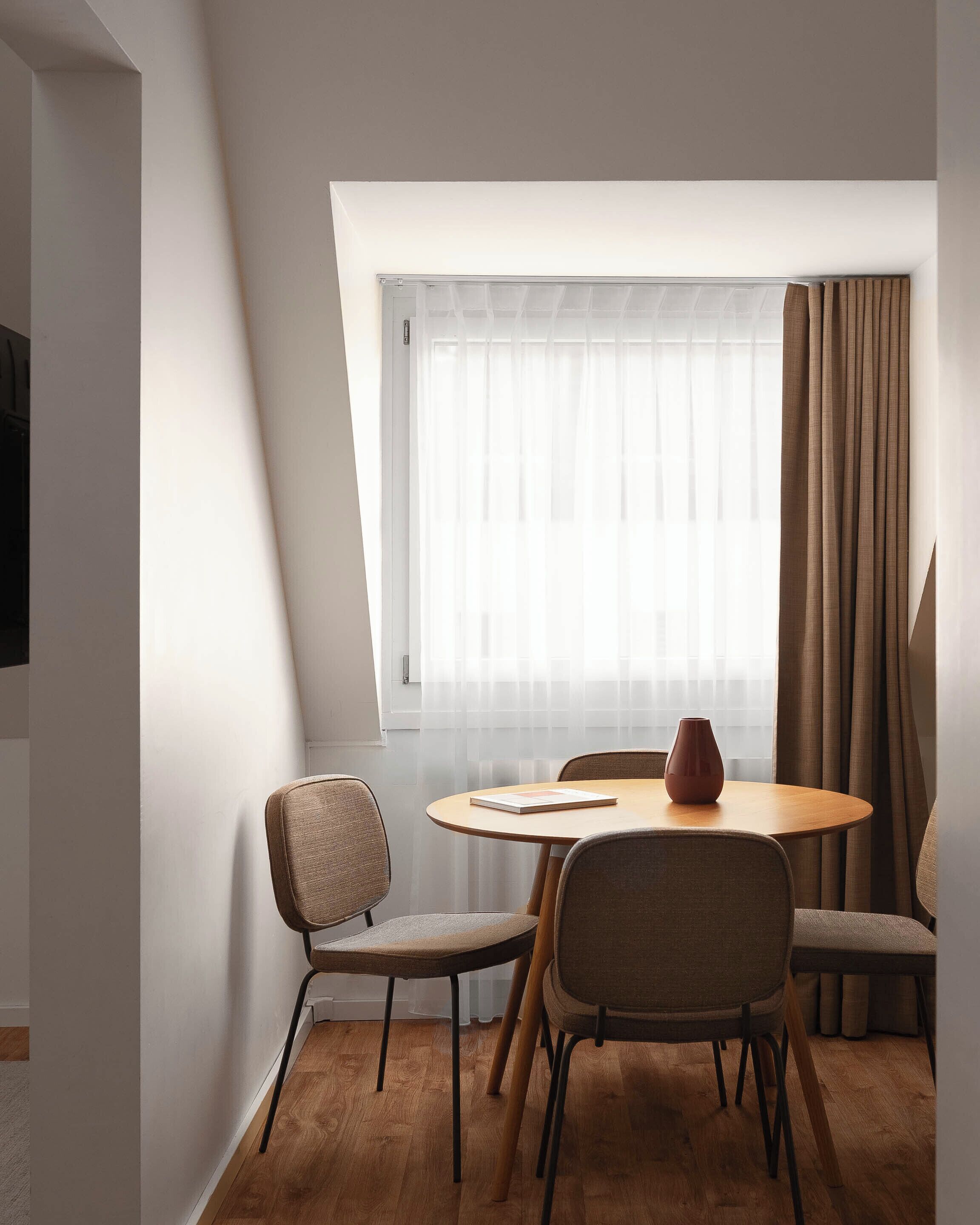 Ein stilvoller Essbereich mit einem runden Tisch, vier Stühlen, einer Vase und einem Fenster mit transparenten Vorhängen.