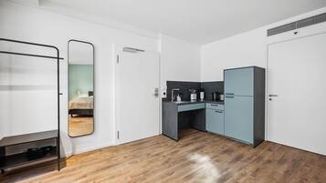 A modern, compact room with a wooden floor, a bed, a desk with a chair, a wardrobe, and a mirror.