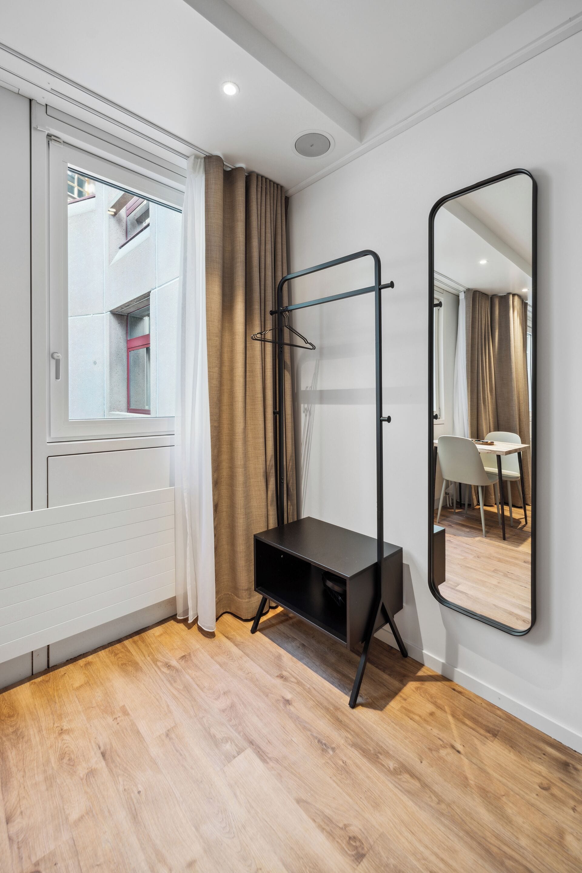 A modern room with a wooden floor, a black desk, a mirror, and a window with curtains.