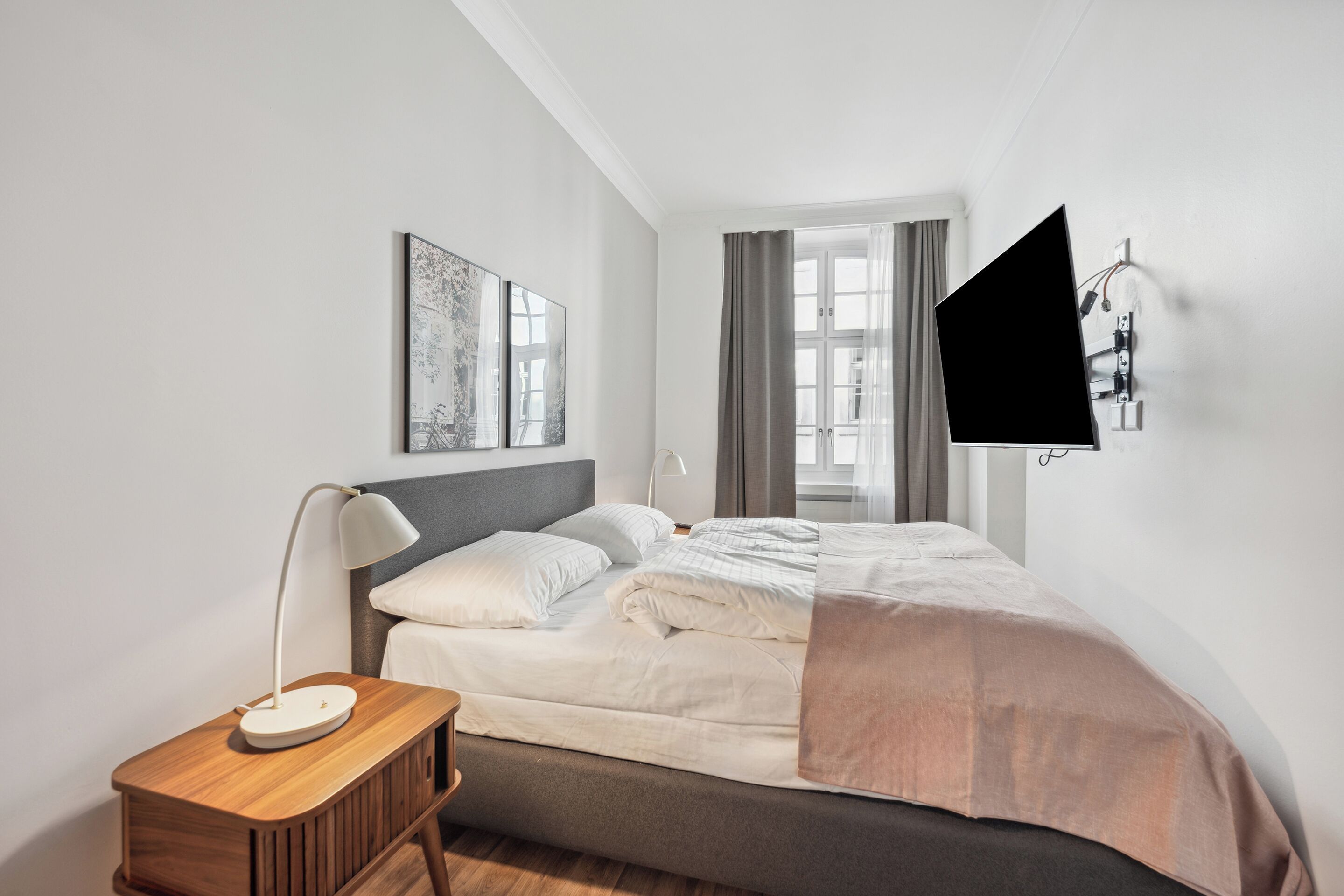 A modern bedroom with a bed, bedside table, lamp, artwork, and a wall-mounted TV.