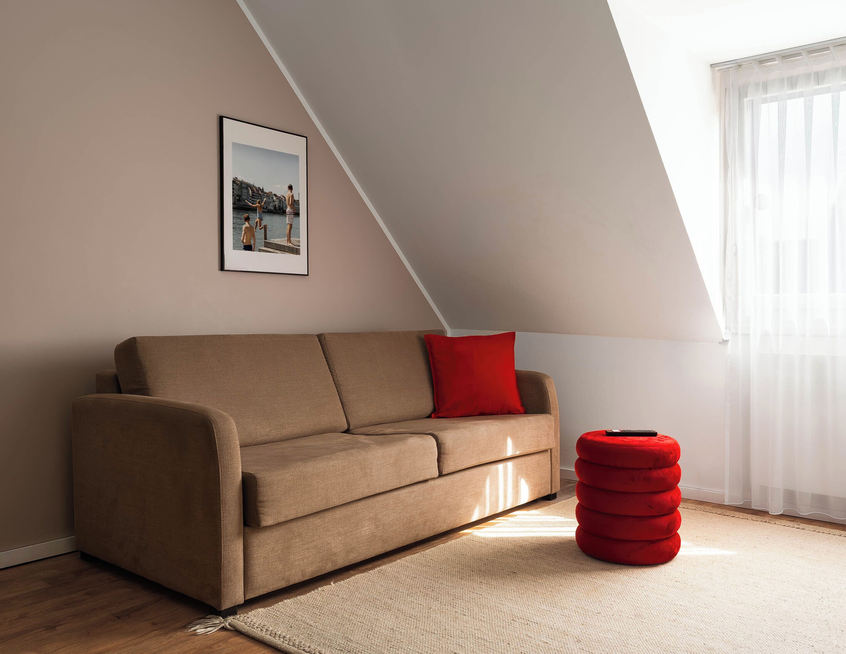 Un salon moderne avec un canapé, un coussin rouge, un tableau encadré et un pouf rouge.