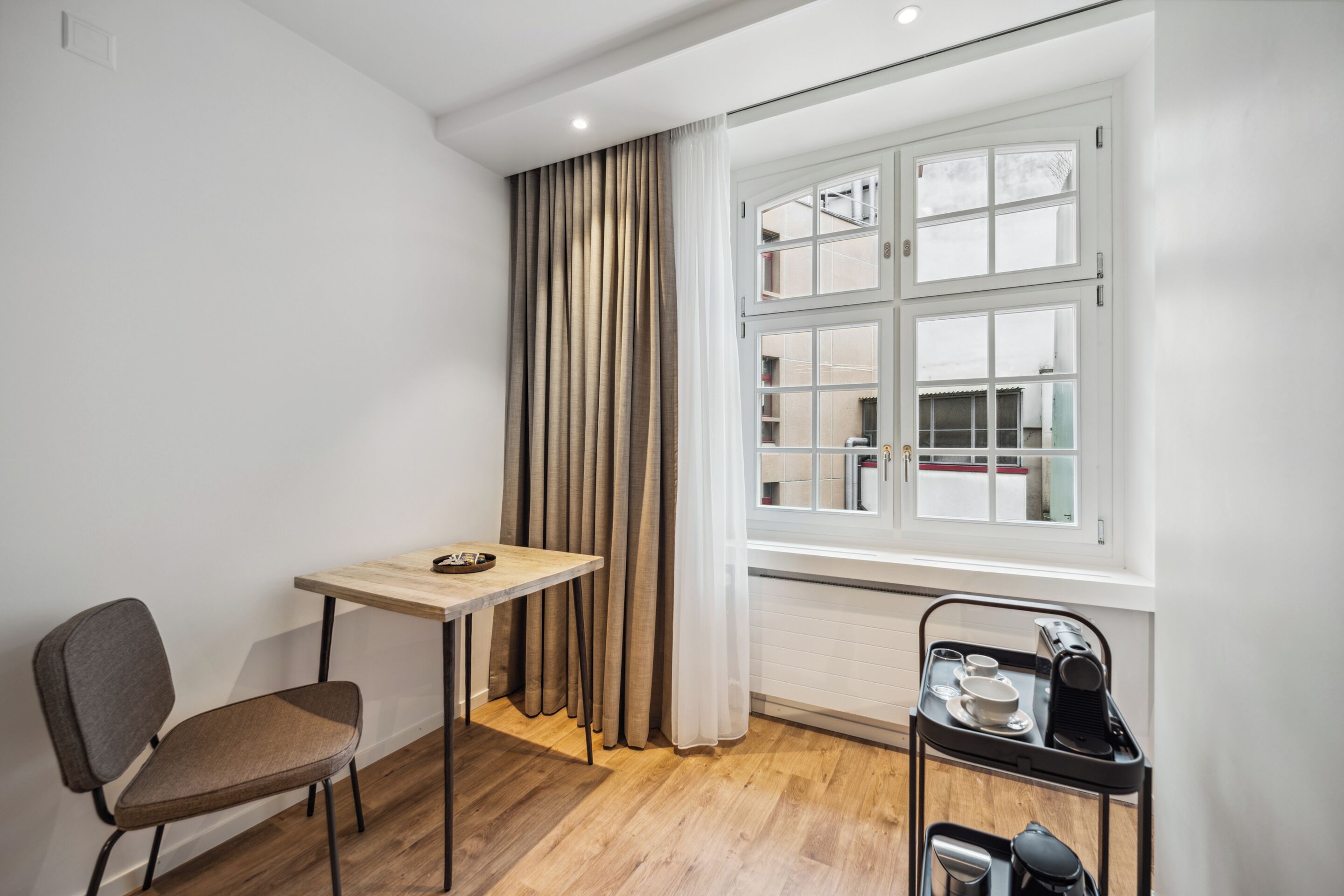 A small, bright room with a wooden floor, a single chair, a table with a tray, and a window with curtains.