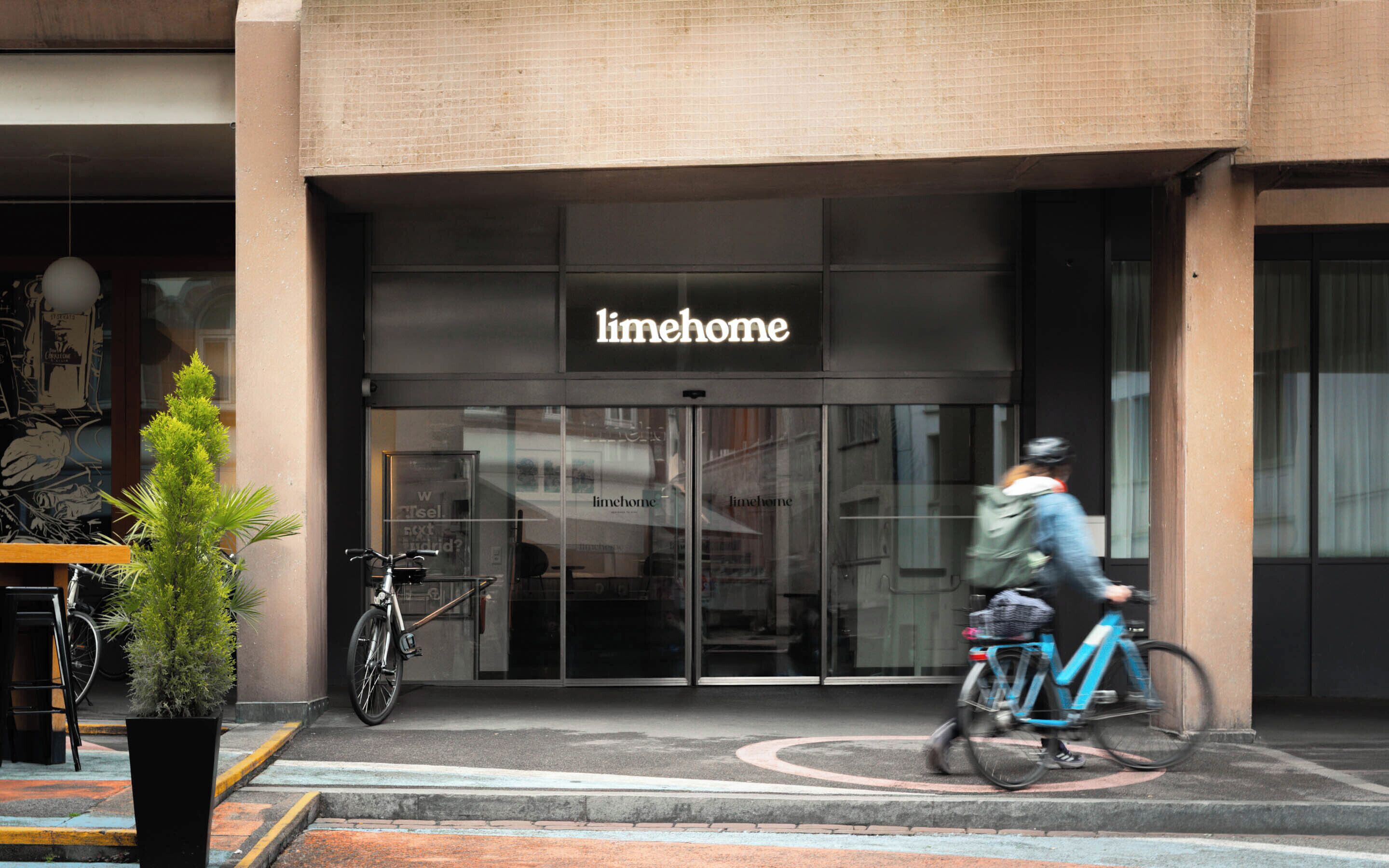 Ein Gebäude mit einem 'limehome'-Schild, ein Fahrrad vor der Tür und ein Pflanzgefäß mit Grünpflanzen.