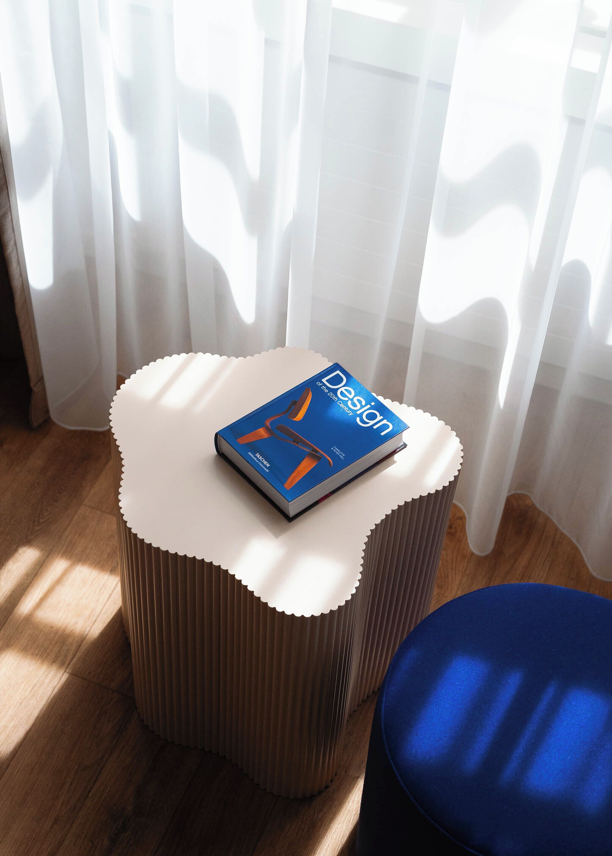 A small round table with a book titled 'Design' on it, placed in a room with wooden flooring and a window with sheer curtains.