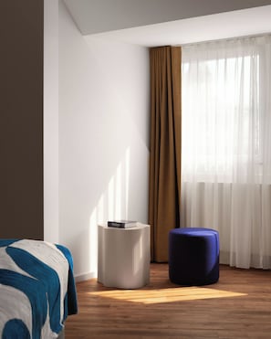 Moderni hotellihuone, jossa on sänky, pyöreä sivupöytä, sininen rahi ja suuri ikkuna, jossa on läpikuultavat verhot.