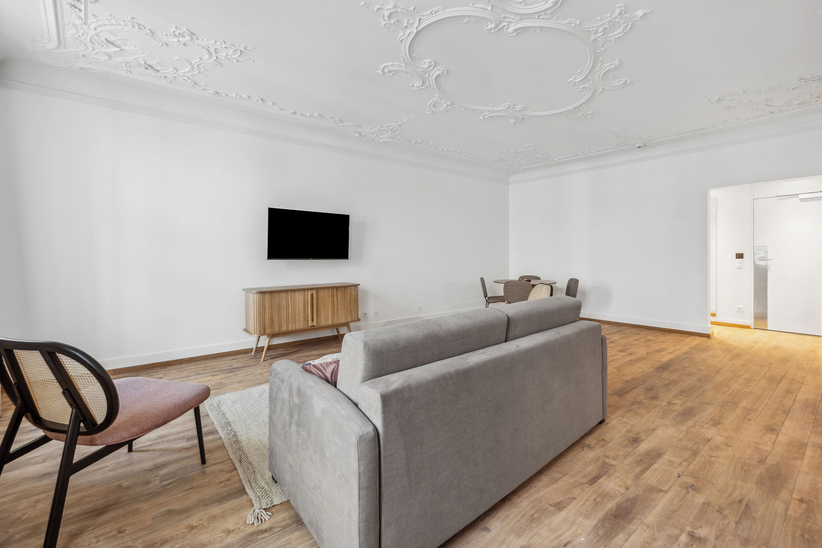 A modern living room with a grey sofa, wooden flooring, a flat-screen TV mounted on the wall, and a small wooden side table.
