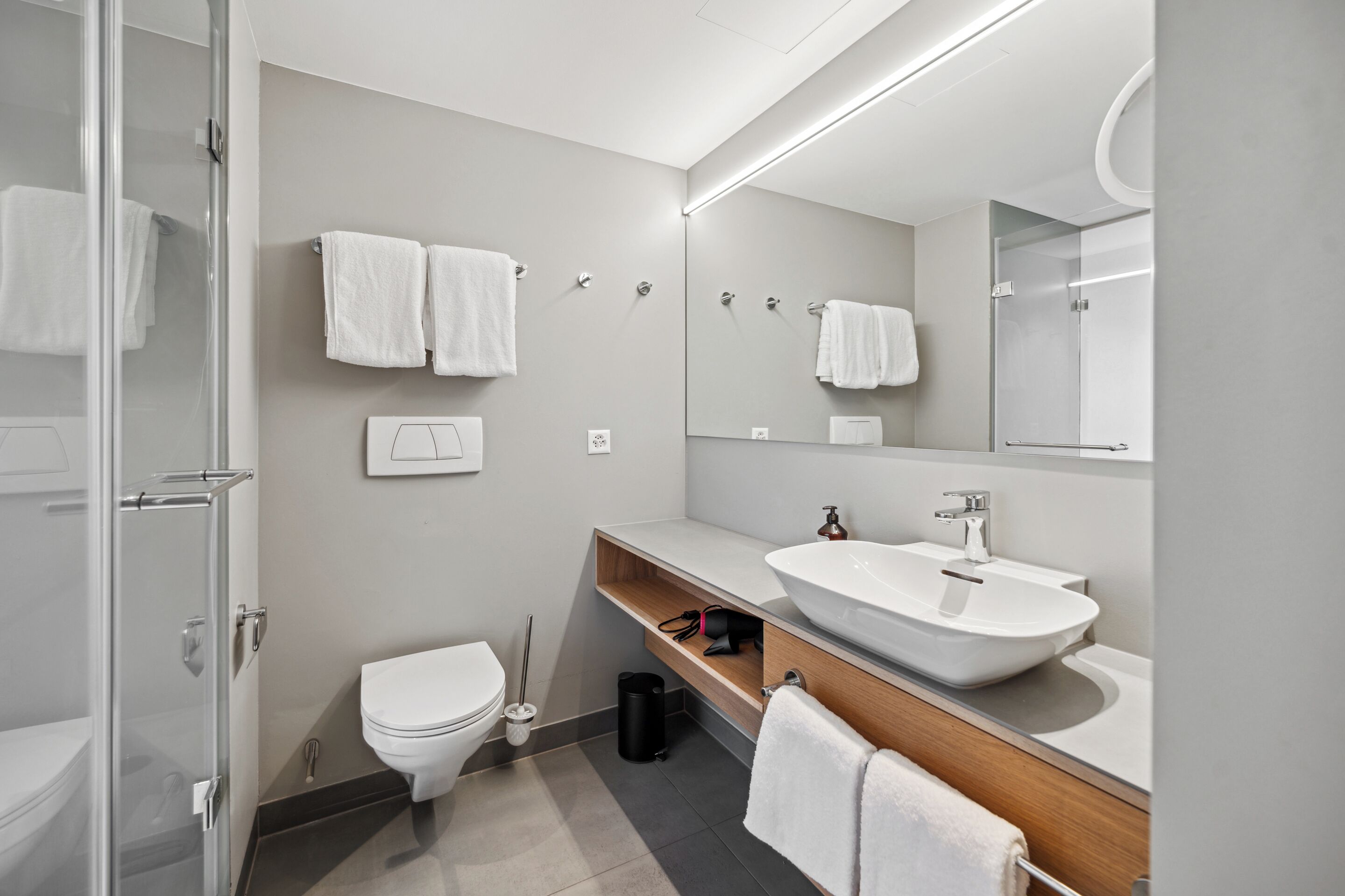 Une salle de bain moderne avec une douche à l’italienne en verre, des toilettes blanches, un grand miroir et un meuble-lavabo en bois avec un vasque.