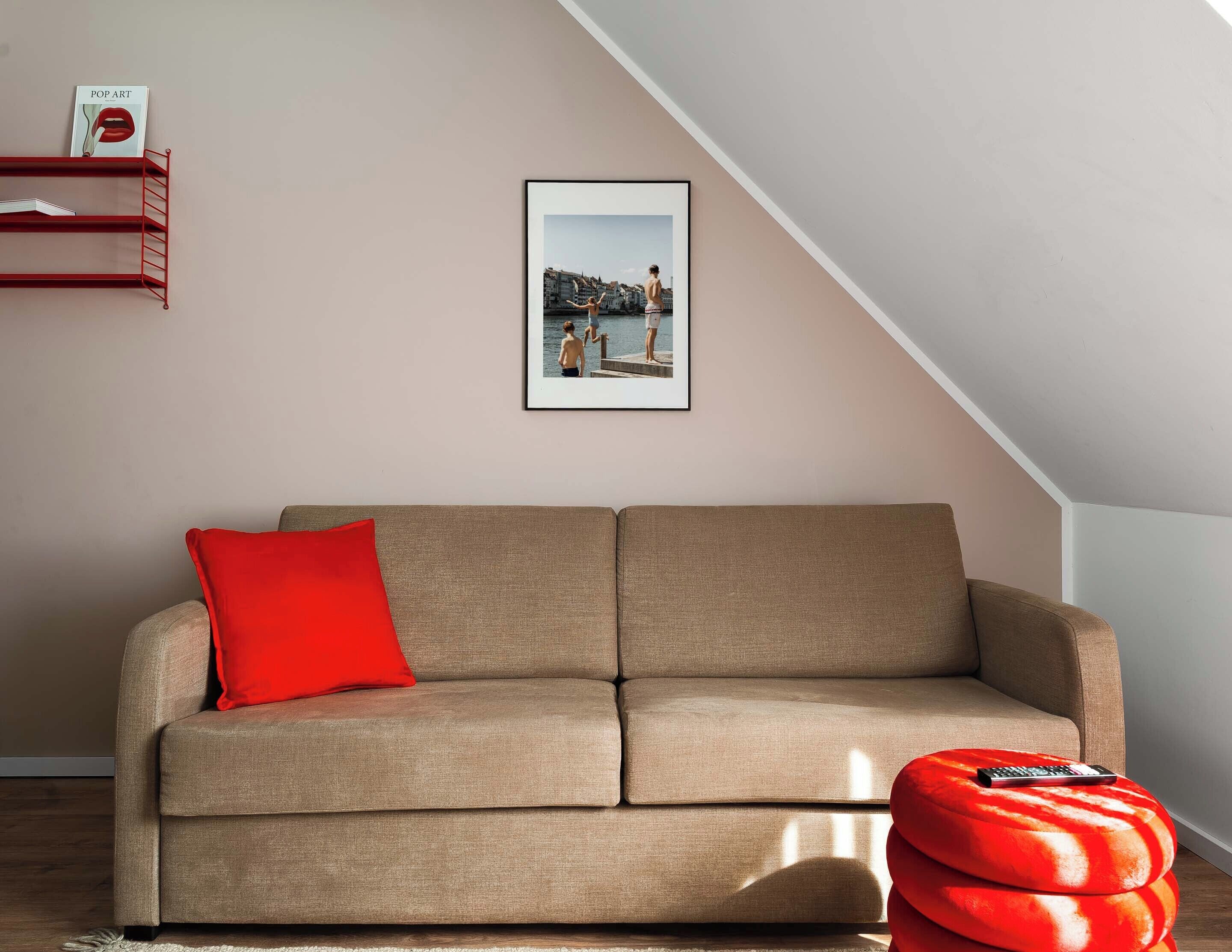 Una sala de estar con un sofá color beige, un cojín rojo, un reposapiés rojo, un cuadro enmarcado en la pared y una estantería de metal rojo.
