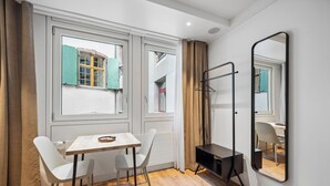 A room with a wooden table and two chairs, a black TV stand, a mirror, and a window with curtains. - Limehome Basel Town Hall (Basel)