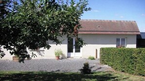 A single-story house with a sloped roof, a front door, and a small front yard with a hedge and a tree. - Apple blossoms (Les Moutiers-en-Cinglais)
