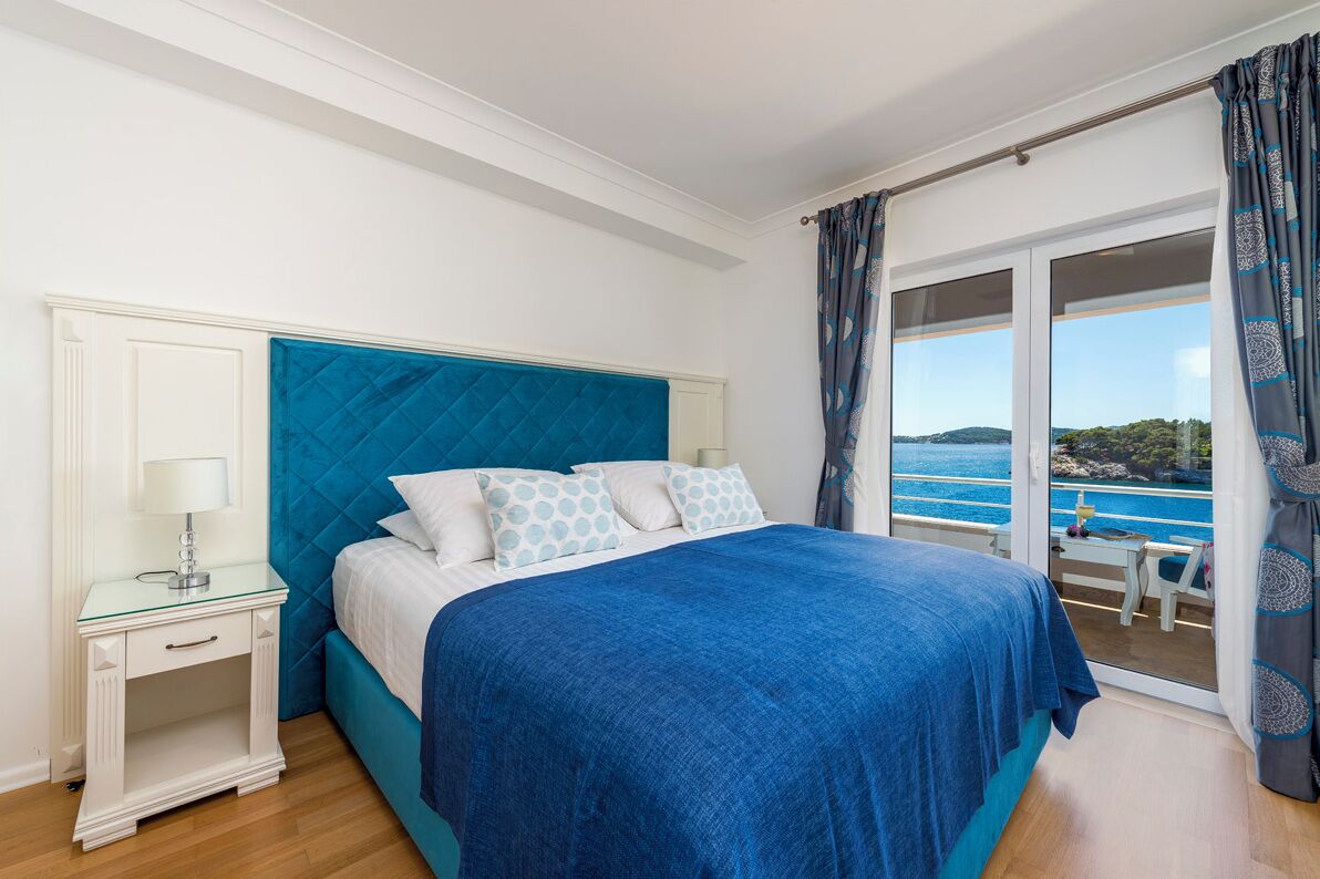 A bedroom with a large bed, a nightstand, a window with a view of the sea, and a balcony with curtains.
