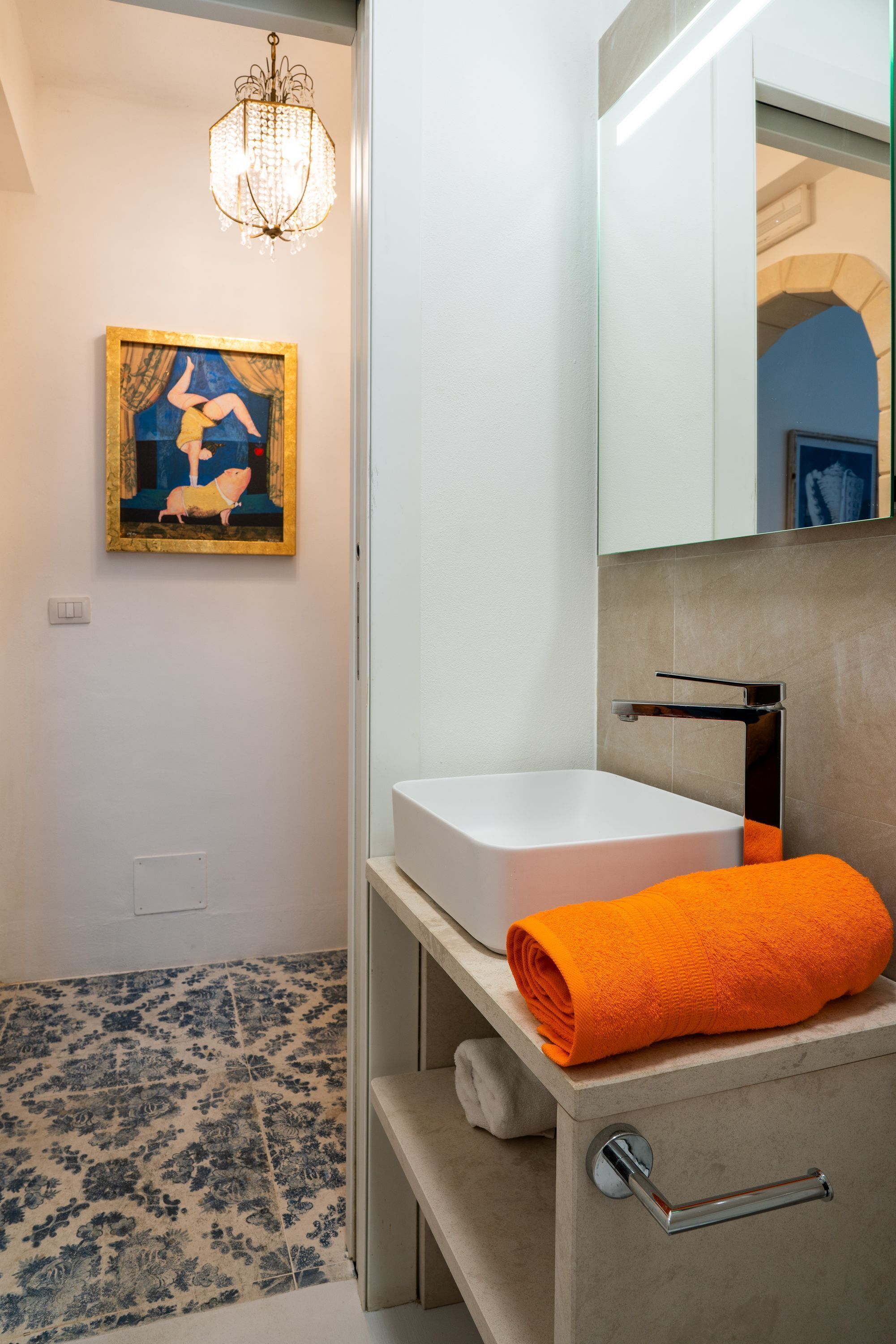 Une salle de bain moderne avec un lavabo blanc, une serviette orange, un lustre et une œuvre d’art encadrée accrochée au mur.