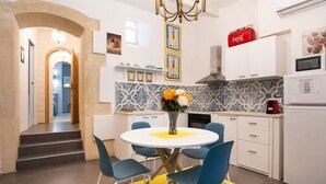 A modern kitchen with white cabinets, a microwave, and a red Coca-Cola cooler. There is a dining area with blue chairs and a yellow table mat. - Ortigia Courtyard Apartment (Syracuse)