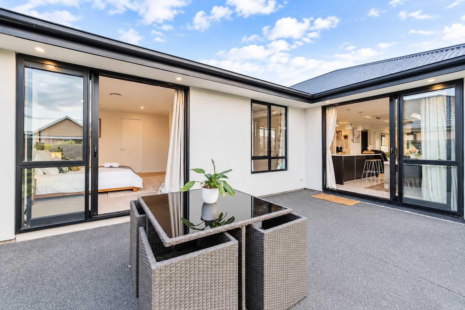 A modern outdoor patio with a glass table and wicker chairs, a bedroom with a bed and curtains, and a kitchen area visible through glass doors.