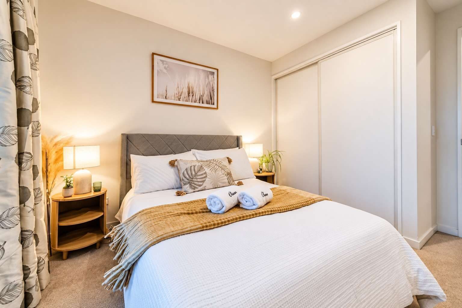 Een netjes opgemaakt bed met kussens en een dekbed, een nachtkastje met een lamp, een ingelijste foto aan de muur, en een gesloten deur.