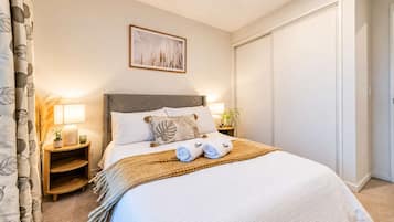Een netjes opgemaakt bed met kussens en een dekbed, een nachtkastje met een lamp, een ingelijste foto aan de muur, en een gesloten deur.