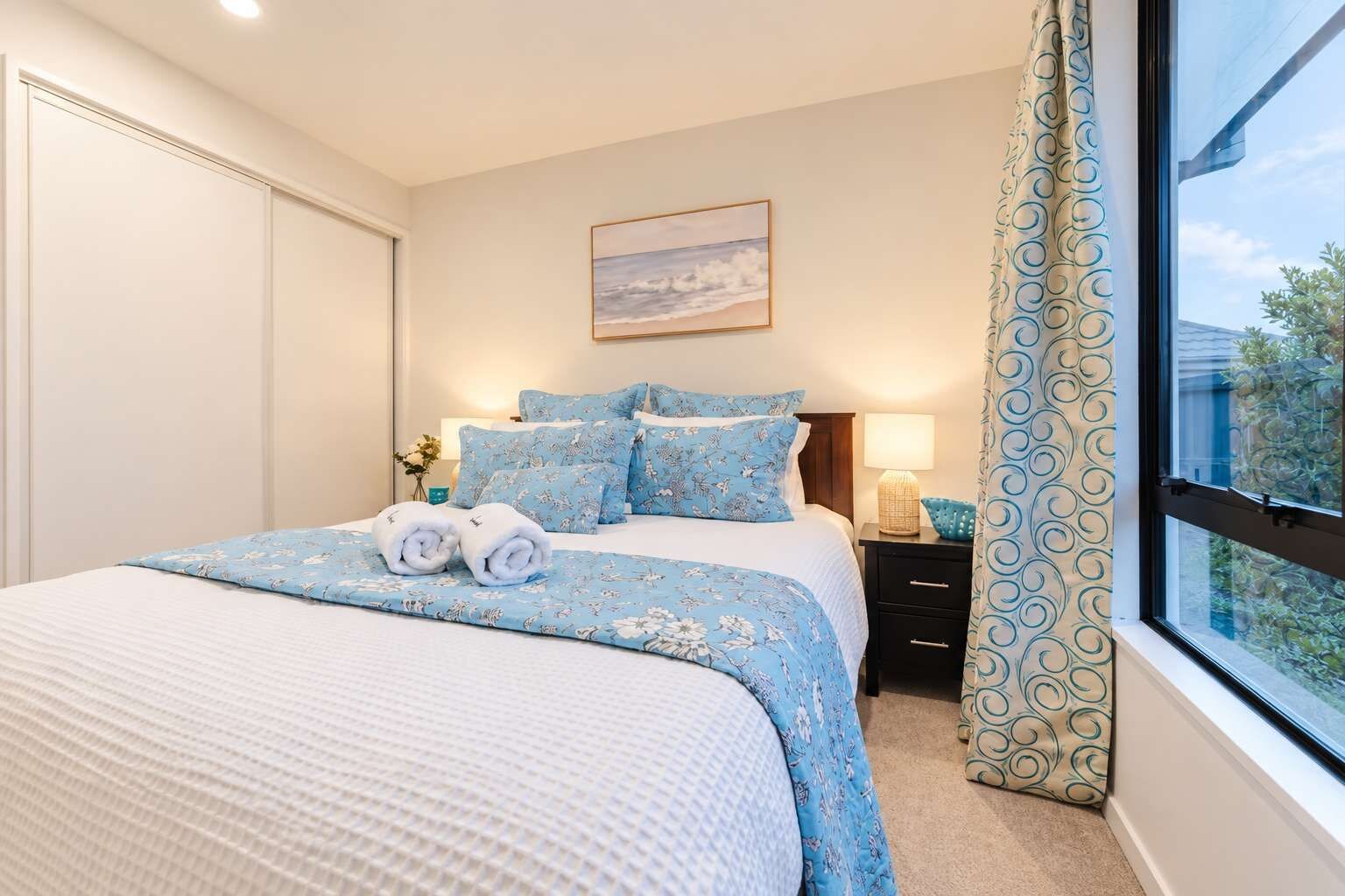 A bedroom with a bed, a nightstand, a lamp, a window with curtains, and a painting on the wall.