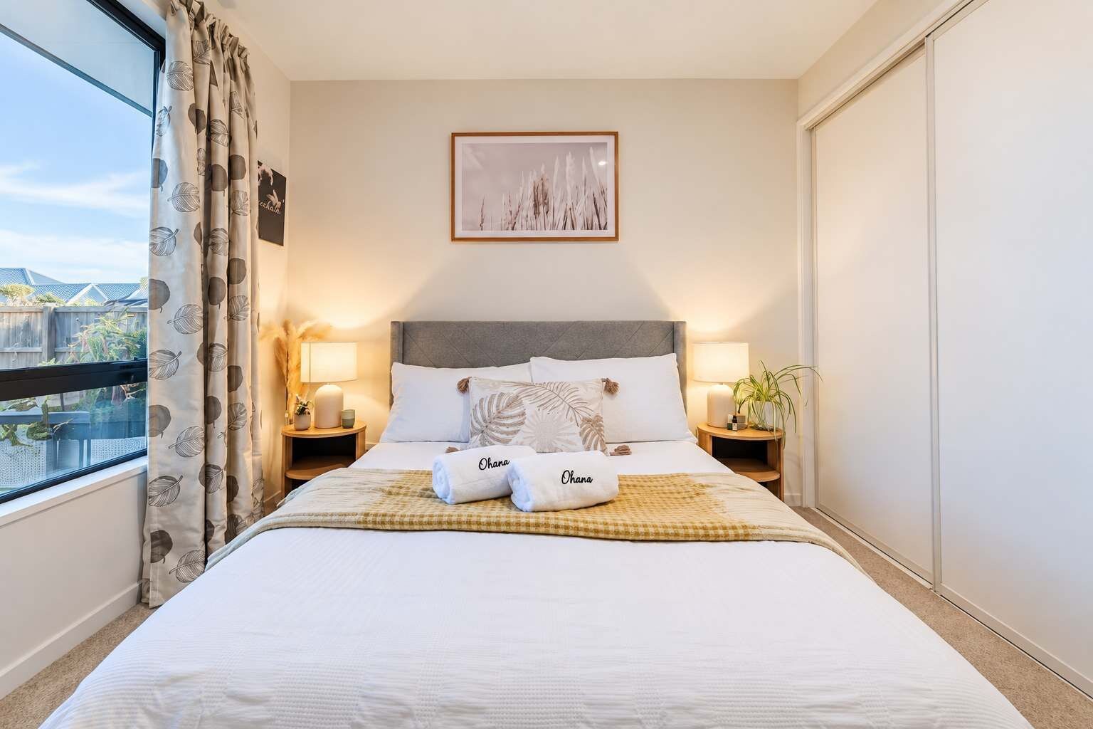 A hotel room with a large bed, two bedside tables, a window with curtains, a framed picture on the wall, and a view of the outside.