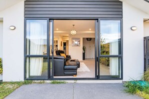 A modern house with a sliding glass door leading to a living area with a sofa and a chandelier. - Sunlit Wigram Home | 4BR + Patio | Sleeps 8 (Christchurch)