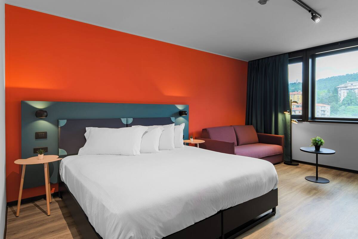 A modern hotel room with a large bed, a sofa, a small table with a plant, and a view of a cityscape through the window.