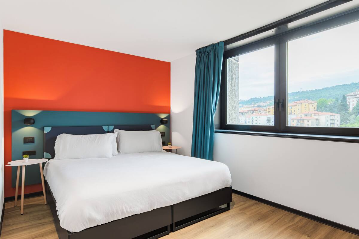 A modern hotel room with a large bed, a view of the city, and a window with curtains.