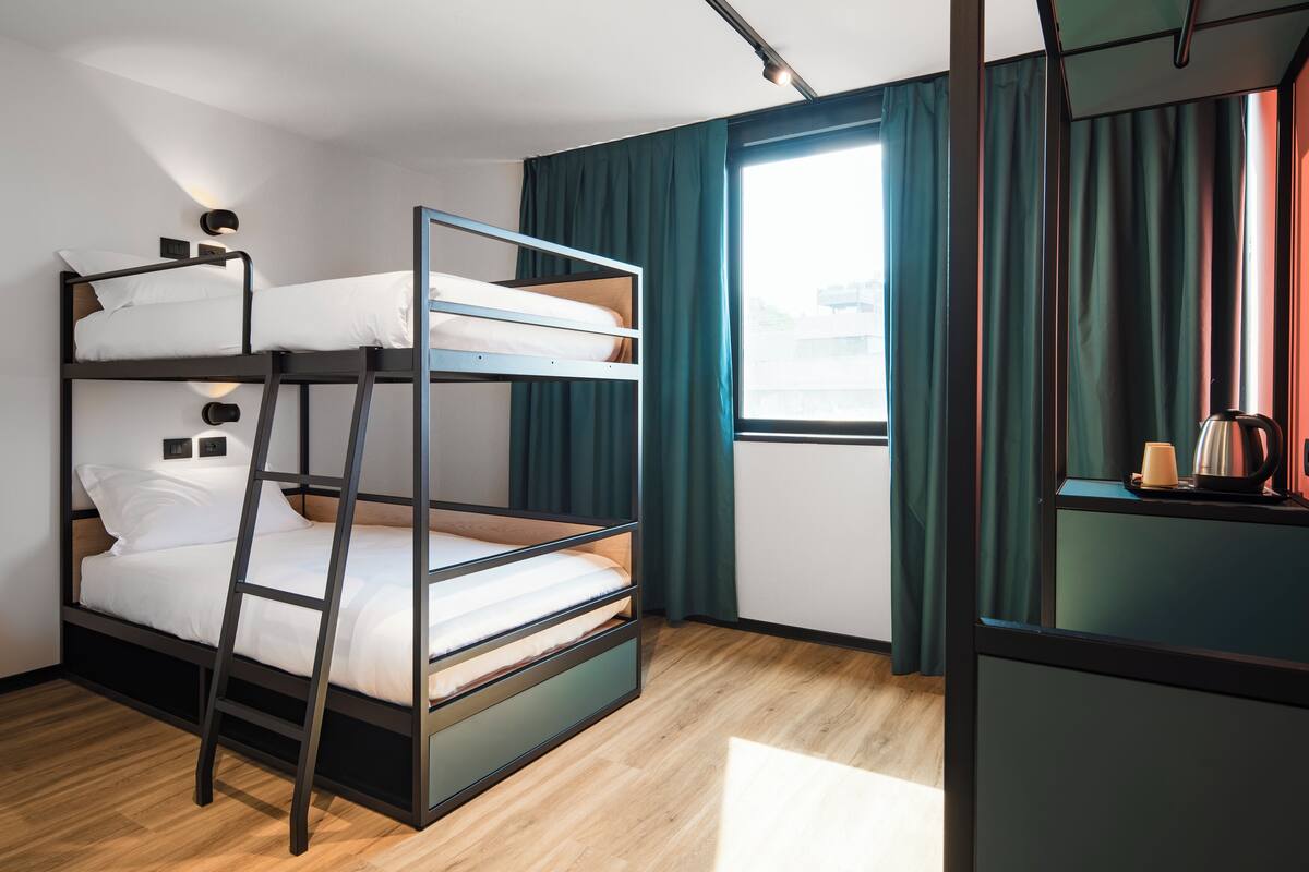 A bunk bed room with a wooden floor, a window with curtains, and a small table with a kettle.
