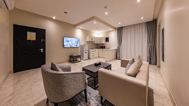 A modern living room with a sofa, armchair, and coffee table, a TV, and a kitchen area with cabinets and a refrigerator. - Towlan Qurtuba 2 Serviced Apartments (Riyadh)