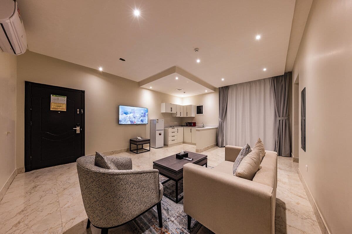 A modern living room with a sofa, armchair, and coffee table, a TV, and a kitchen area with cabinets and a refrigerator.