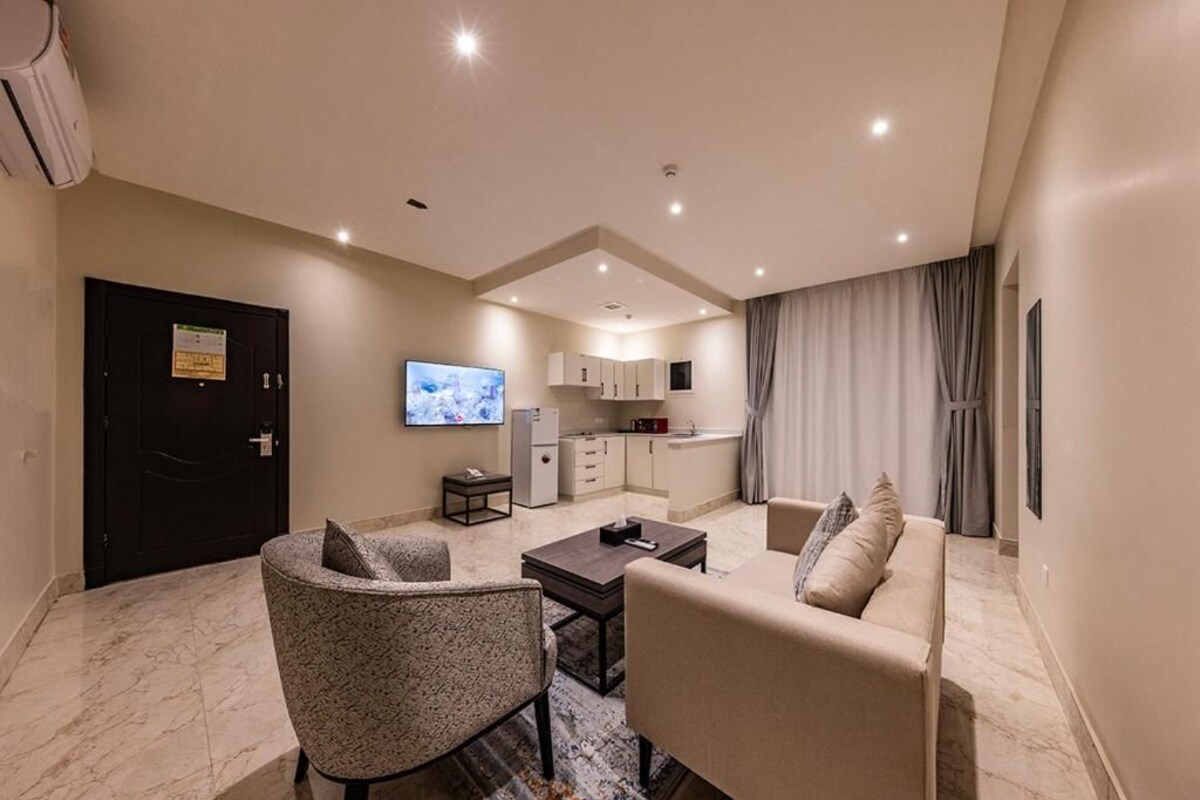 A modern living room with a sofa, armchair, and coffee table. A TV is mounted on the wall, and a kitchen is visible in the background.