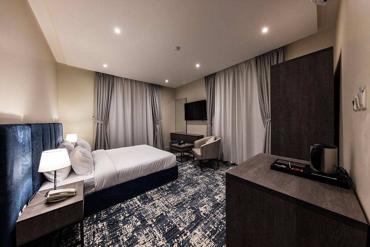 A modern hotel room with a bed, a desk, a chair, a TV, and a minibar.