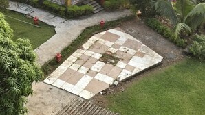 Un edificio pequeño de una sola planta con techo de tejas, rodeado de exuberante vegetación y un área pavimentada con diseño de cuadros.