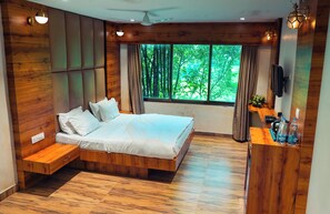 Un dormitorio moderno con cama de madera, un banco empotrado, televisor y un ventanal con vistas a árboles.