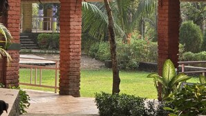 Un espacio al aire libre cubierto con pilares de ladrillo, suelo de concreto y abundante vegetación de fondo.