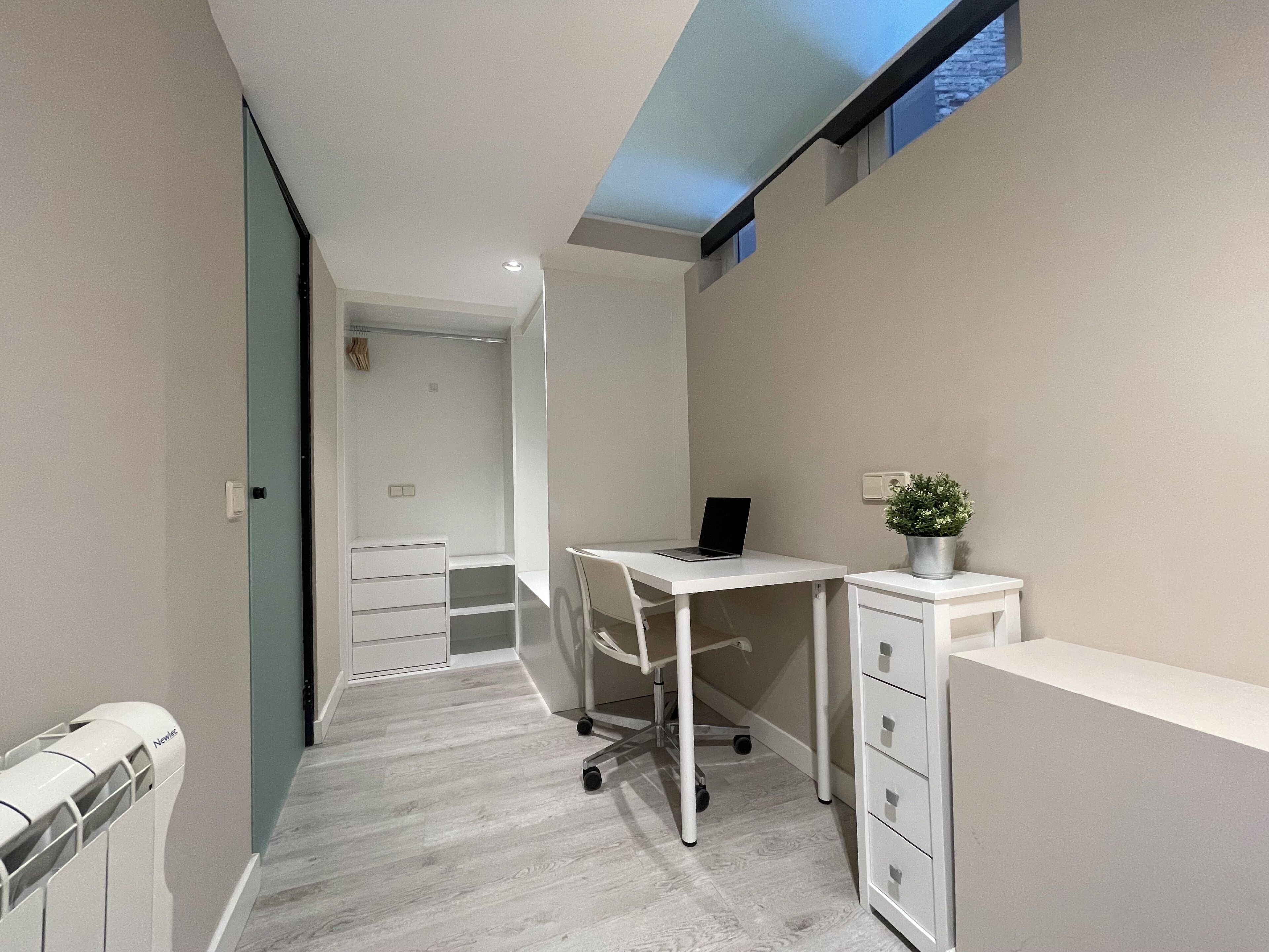 Una habitación moderna y estrecha con un escritorio, una silla, un radiador y una pequeña planta en maceta.